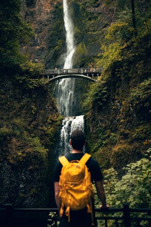 A hiker with a yellow backpack stands before the stunning two-tiered Multnomah Falls waterfall in Oregon.