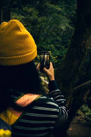 Person in yellow beanie photographing forest waterfall on smartphone among lush green trees.