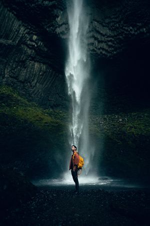 A hiker with a yellow backpack stands in awe beneath a towering waterfall inside a dark dramatic cave.