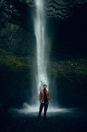A man in an orange jacket stands before a dramatic tall waterfall in a dark rocky gorge.