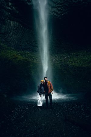 Couple standing together in front of a tall dramatic waterfall in a dark mossy canyon.