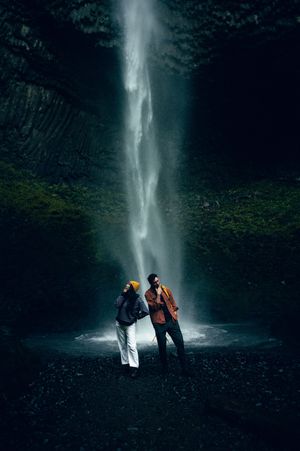 A couple stands together beneath a dramatic waterfall in a dark rocky cave setting.