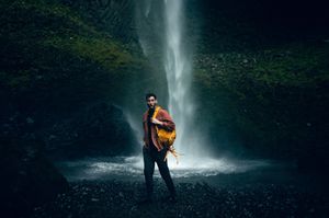 A man with a colorful backpack poses in front of a misty waterfall in a dark cave.
