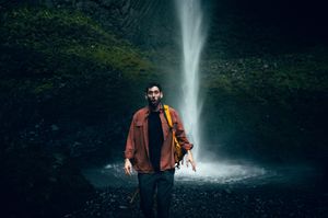 A man in an orange jacket poses in front of a powerful waterfall cascading in a dark rocky grotto.
