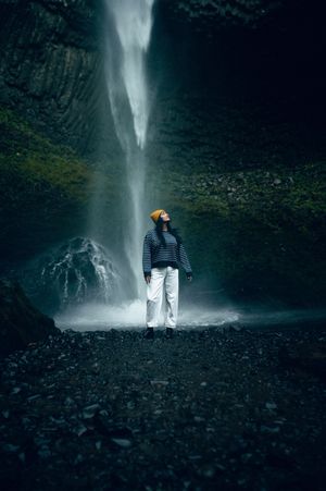 A woman in white pants and yellow beanie stands beneath a dramatic waterfall in a dark rocky cave.