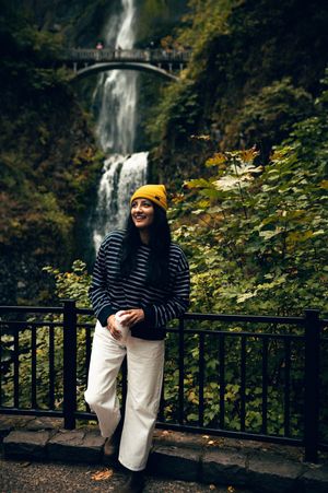 A smiling woman in a yellow beanie stands before Multnomah Falls waterfall in autumn.