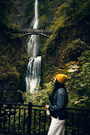 Woman in yellow beanie views Multnomah Falls waterfall with historic bridge in Columbia River Gorge.