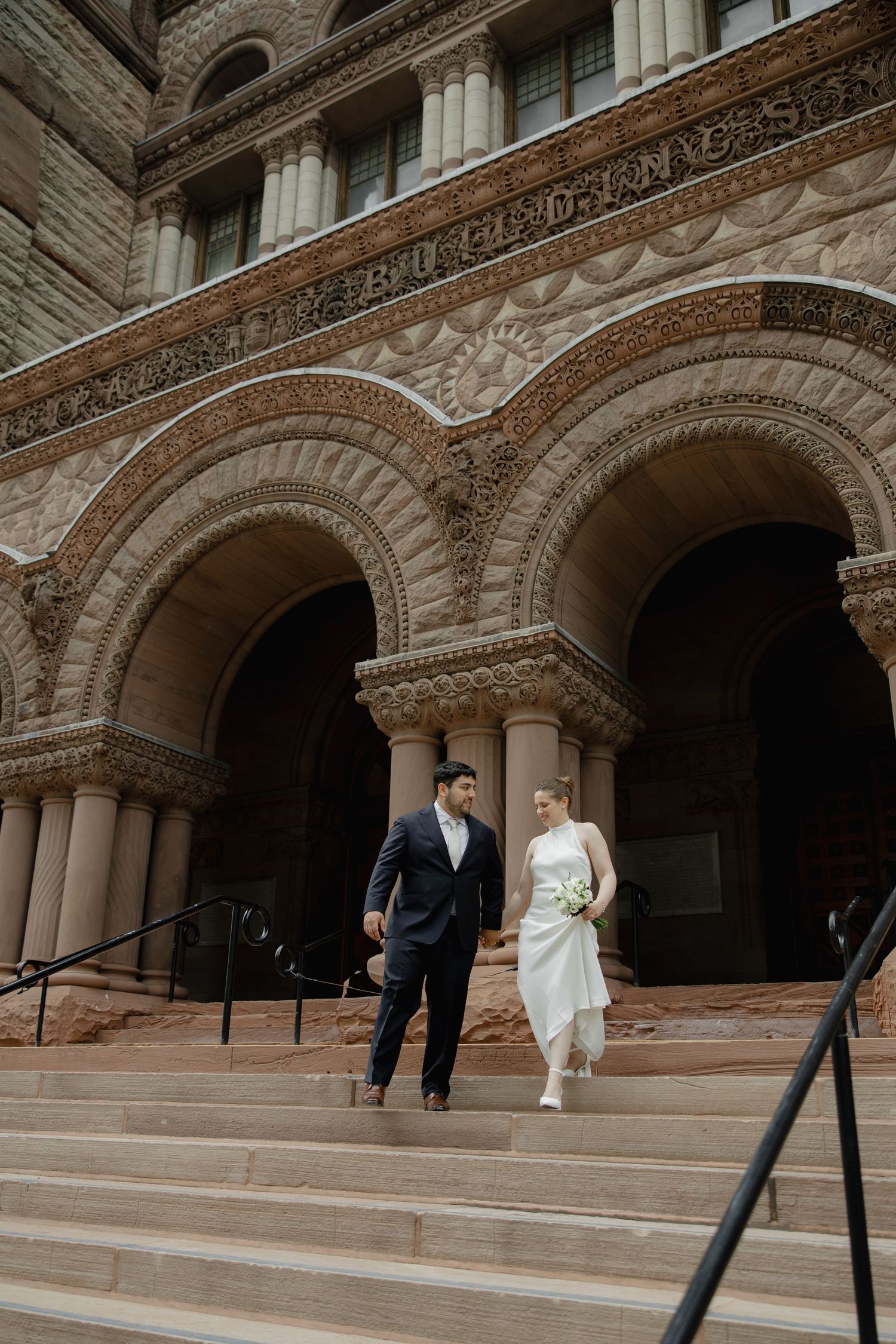Bride and groom walk down stone steps of ornate historic building with arched doorways on their wedding day.