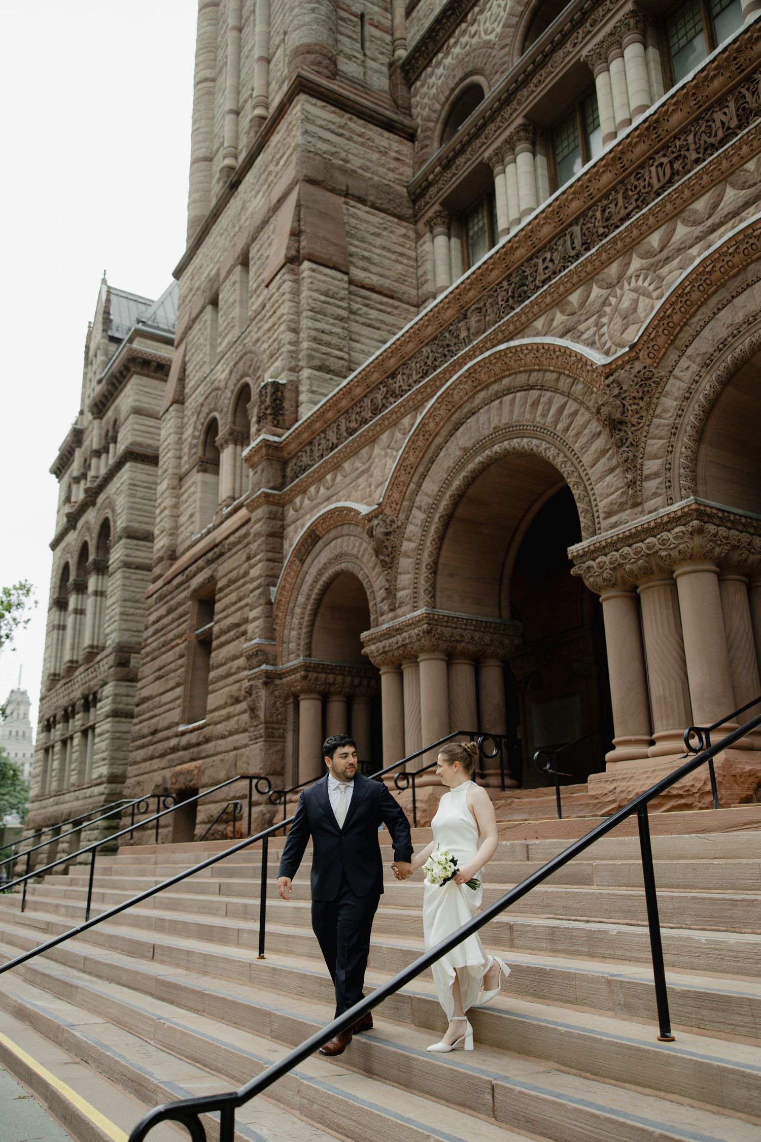 Bride and groom walk down stone steps of ornate historic building, bride holding bouquet in white dress.