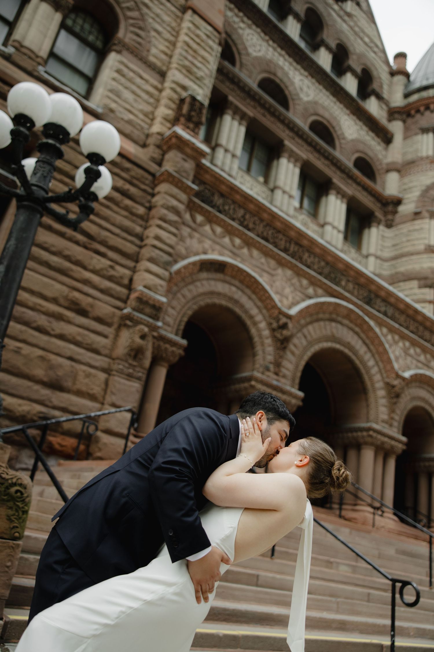 Bride and groom share a romantic dip kiss on the steps of a grand historic stone building on their wedding day.