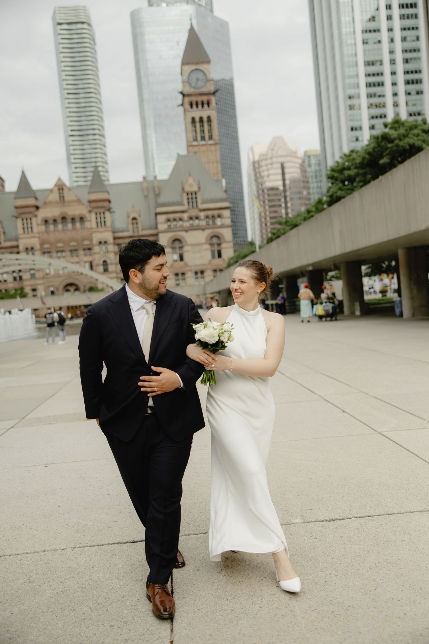 Bride and groom walk together at Nathan Phillips Square with Toronto's Old City Hall in the background.