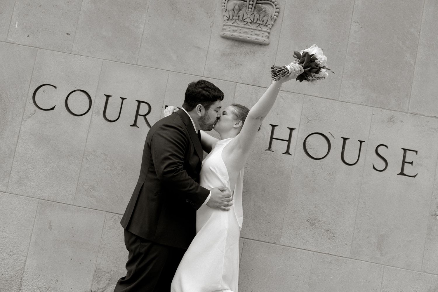 Newlyweds kiss and celebrate outside a courthouse in a classic black and white wedding photo.