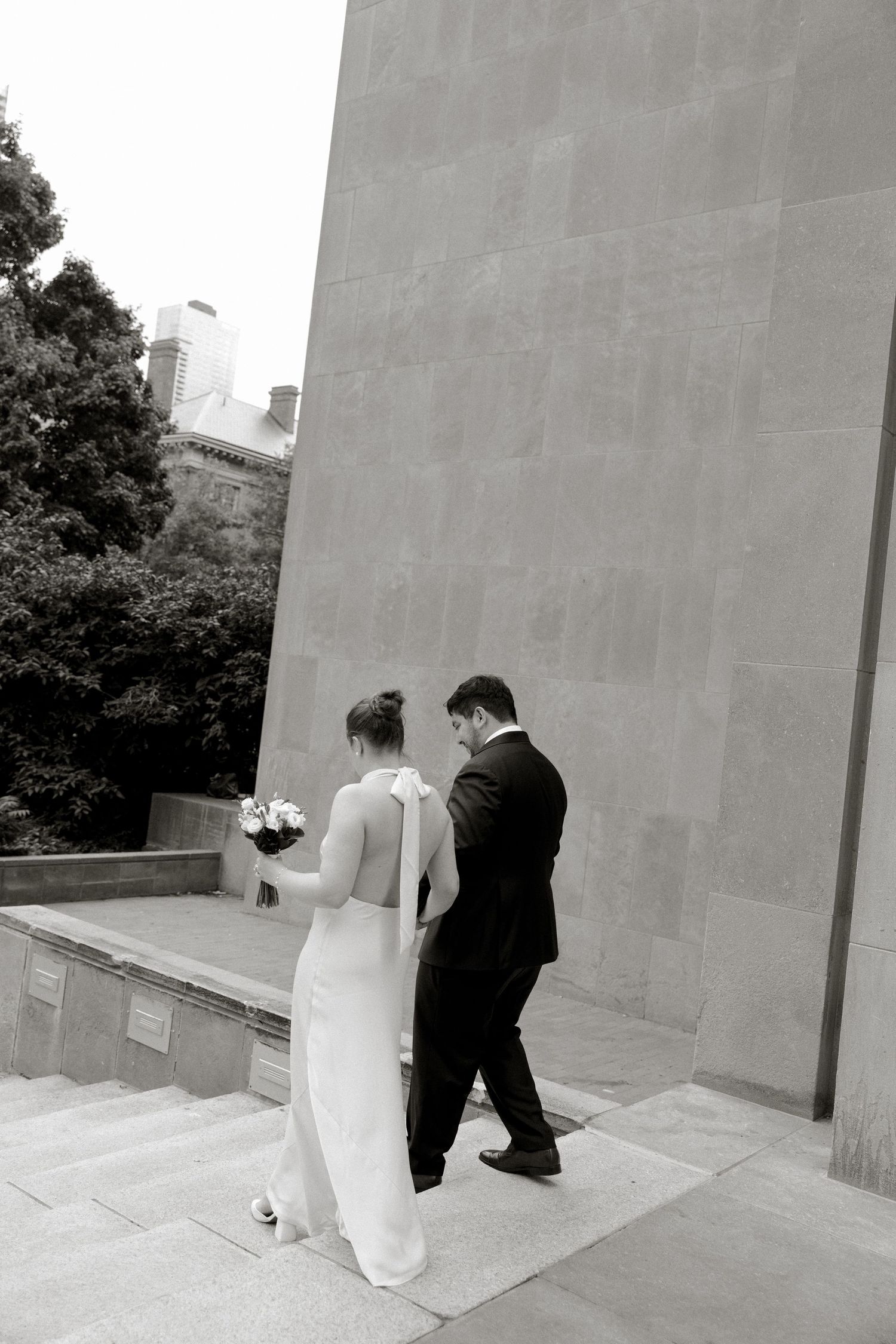 Black and white photo of a bride holding bouquet and groom in suit walking up stone steps near a large wall.