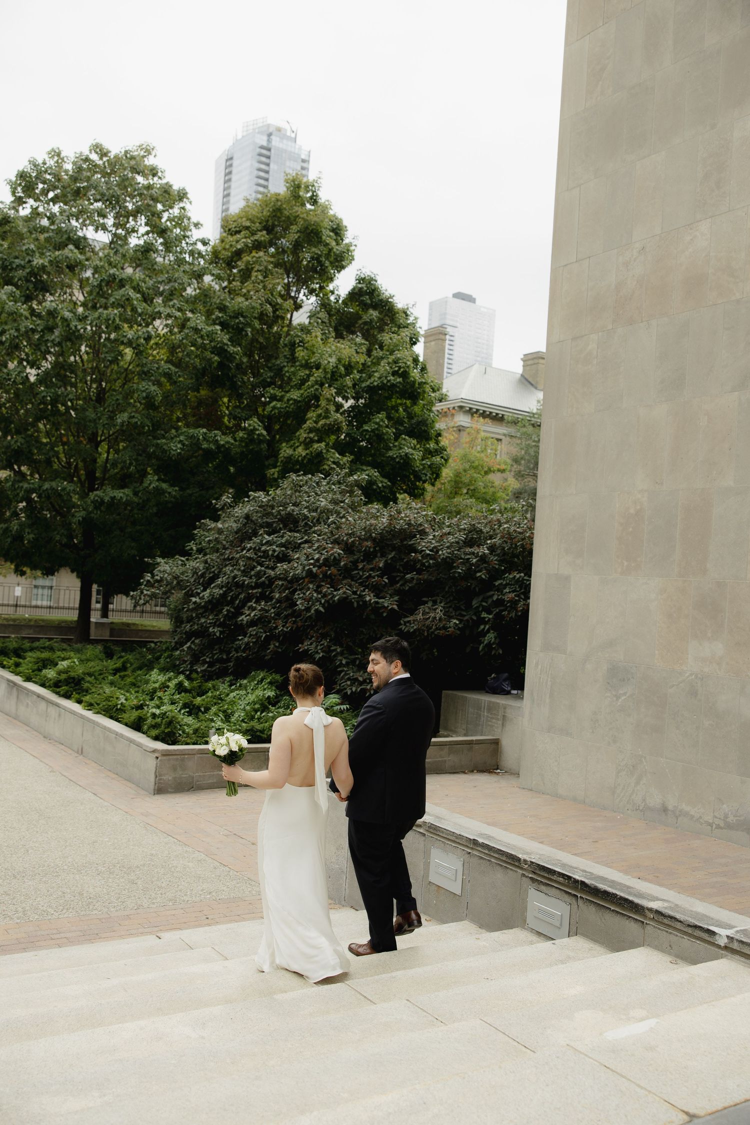 Bride and groom walk up stone steps outside a modern building with lush green trees and city skyline backdrop.