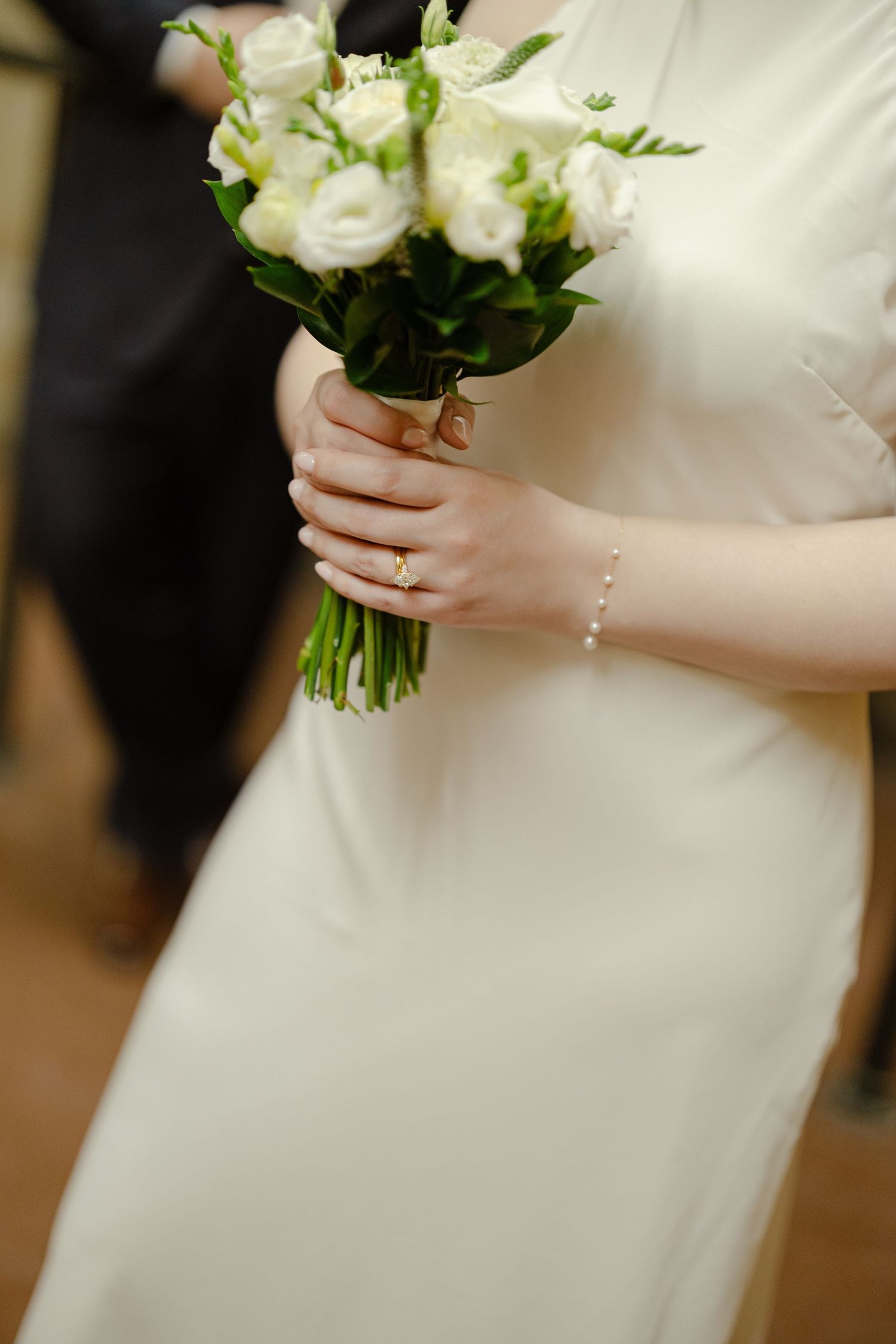 Bride in white dress holding elegant bouquet of white roses and greenery, wearing gold ring and pearl bracelet.