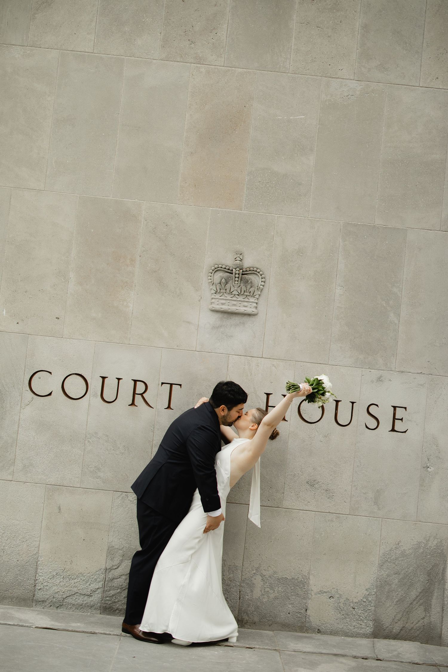 Bride and groom share a romantic dip kiss outside a courthouse, bride holding bouquet aloft in wedding attire.