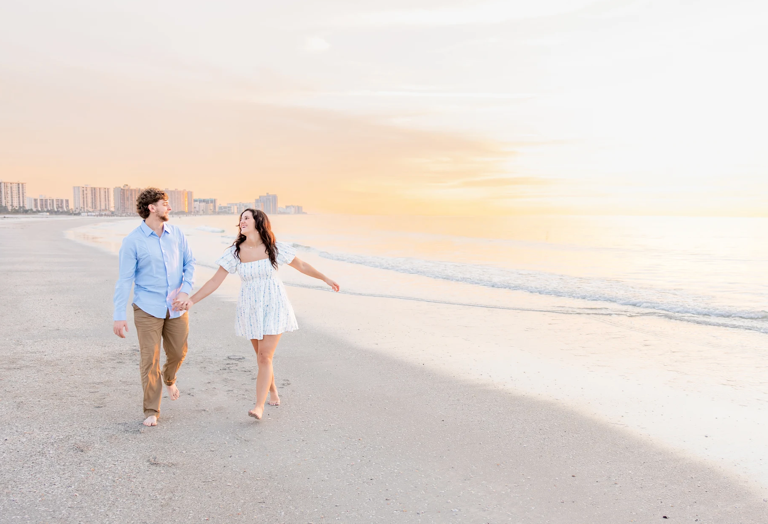 Couple walks hand-in-hand on a beach at sunset, woman in white dress, man in blue shirt, ocean waves behind them.