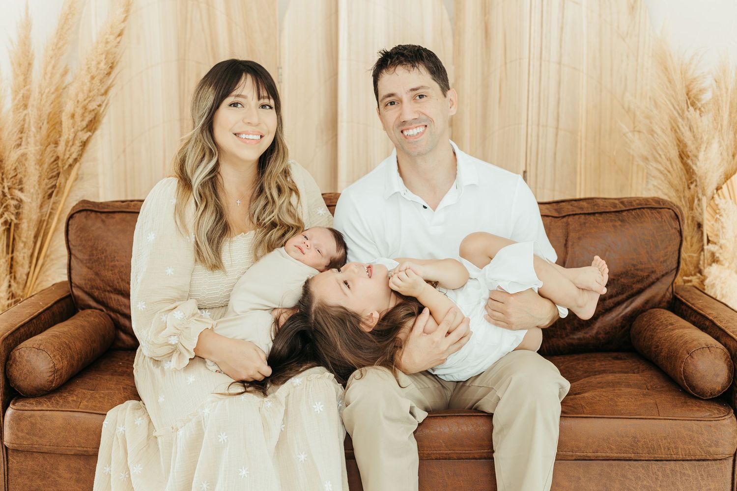family sits on a brown leather couch, parents holding kids in a warm, neutral-toned studio during lifestyle newborn session in kemah TX