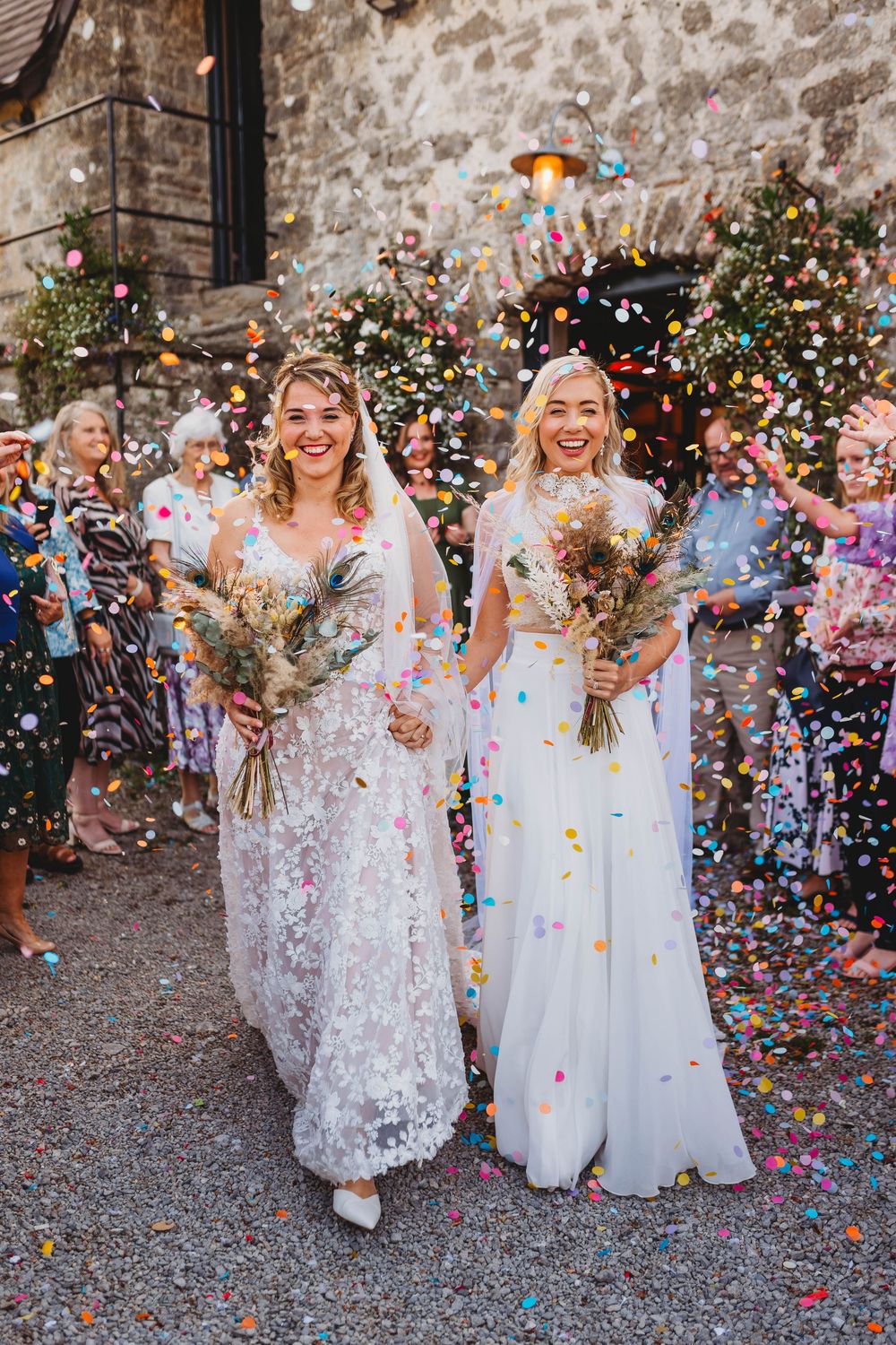 Two smiling brides in white lace wedding gowns hold dried flower bouquets as colorful confetti fills the air outside a stone building.