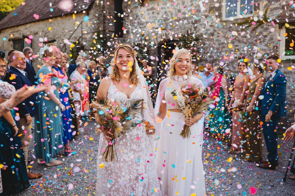 Two brides in white gowns hold bouquets and smile as wedding guests throw colorful confetti around them outside a rustic stone building.
