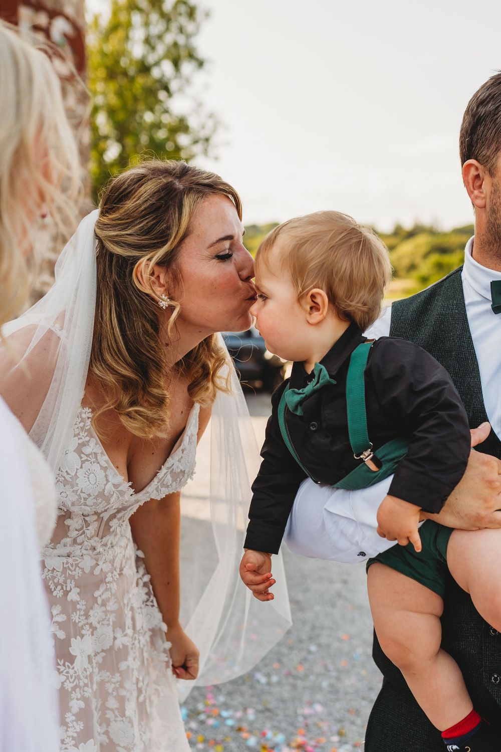 Bride in a lace wedding gown and veil kisses a toddler in a green and black outfit held by a man in a vest at a wedding.
