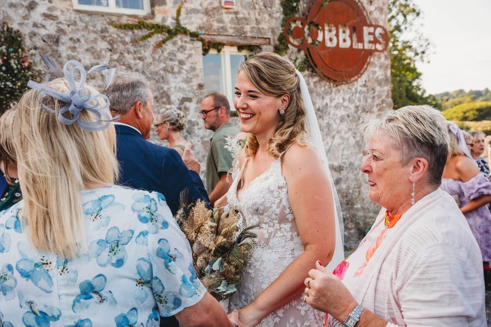 Smiling bride in a lace wedding gown holding a bouquet as guests chat outside a rustic stone venue on a sunny day.