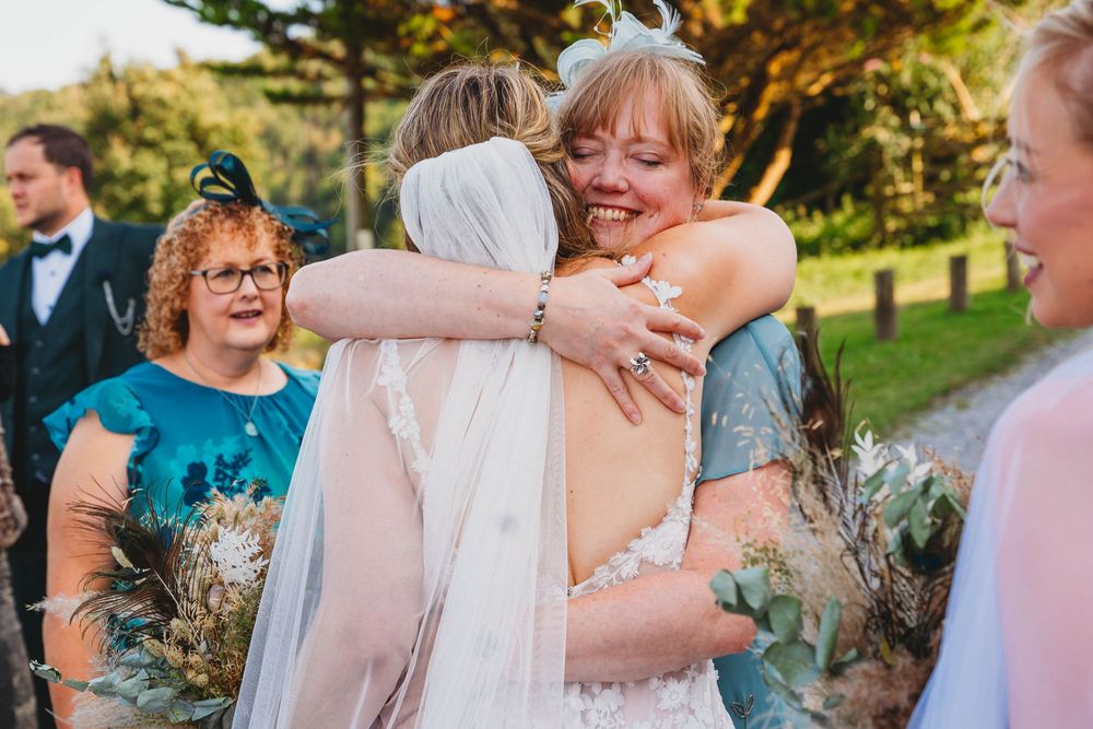A bride in a white wedding dress and veil is embraced warmly by a smiling guest at an outdoor wedding, with other guests nearby.