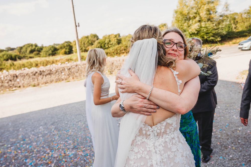 A bride in a lace wedding gown and veil shares a warm embrace with a smiling woman in glasses at an outdoor countryside wedding.
