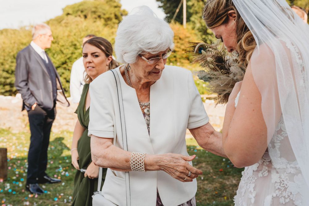 Elderly woman in a white cardigan holds a bride's hand during an outdoor wedding as guests in green dresses and suits look on.