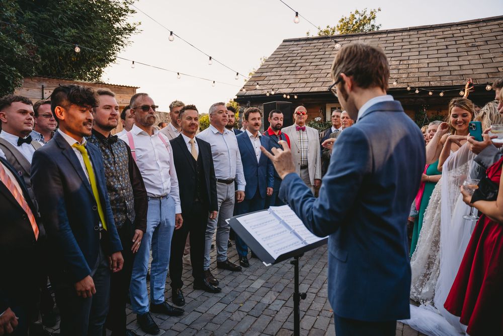 Man in a blue suit conducts a group of smiling men singing at an outdoor wedding as guests look on.