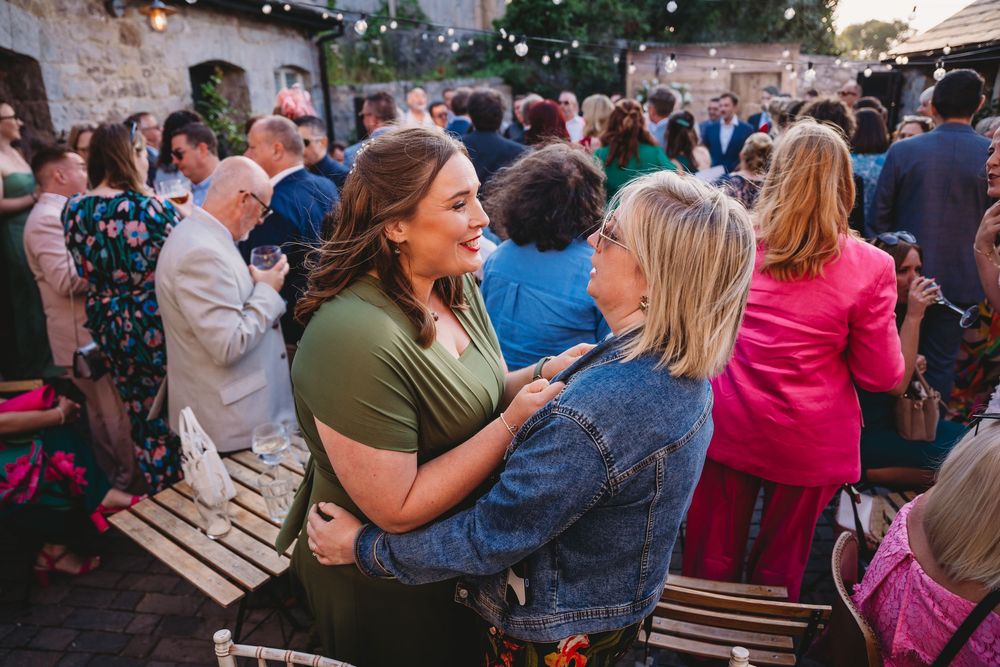 Two women embrace and smile at a lively outdoor evening gathering as guests chat around wooden tables under string lights.