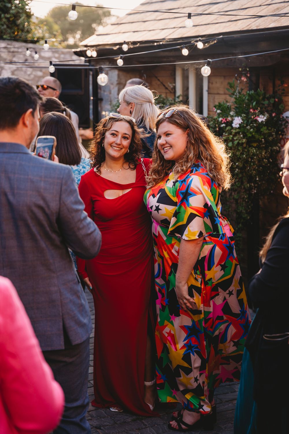 Two smiling women in a red dress and a colorful star-print maxi dress stand together at an outdoor evening gathering under string lights.