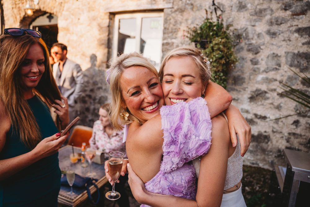 Two women share a joyful hug and smile at an outdoor gathering by a stone building while guests chat and hold drinks nearby.