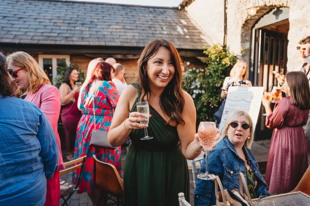 Smiling woman in a green dress holds a champagne glass at a lively outdoor garden party with guests mingling nearby.