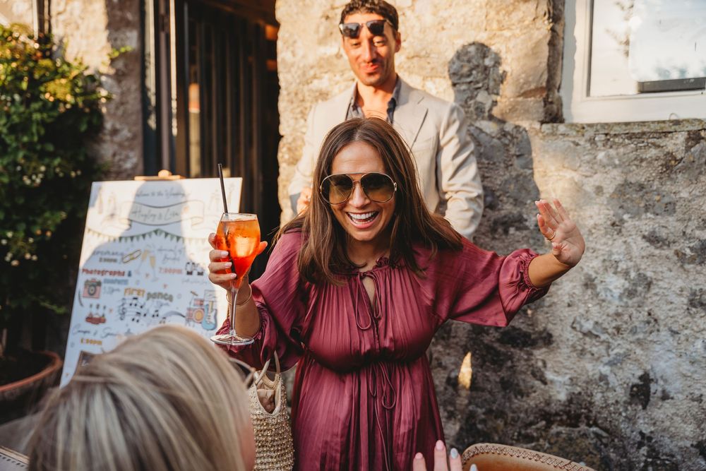 Smiling woman in sunglasses and a burgundy dress holds an orange spritz cocktail and waves at a festive outdoor gathering.