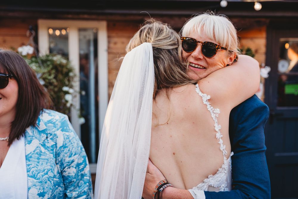 Bride in a lace-back wedding dress and veil hugs a smiling guest in sunglasses while another guest in a blue floral jacket looks on.