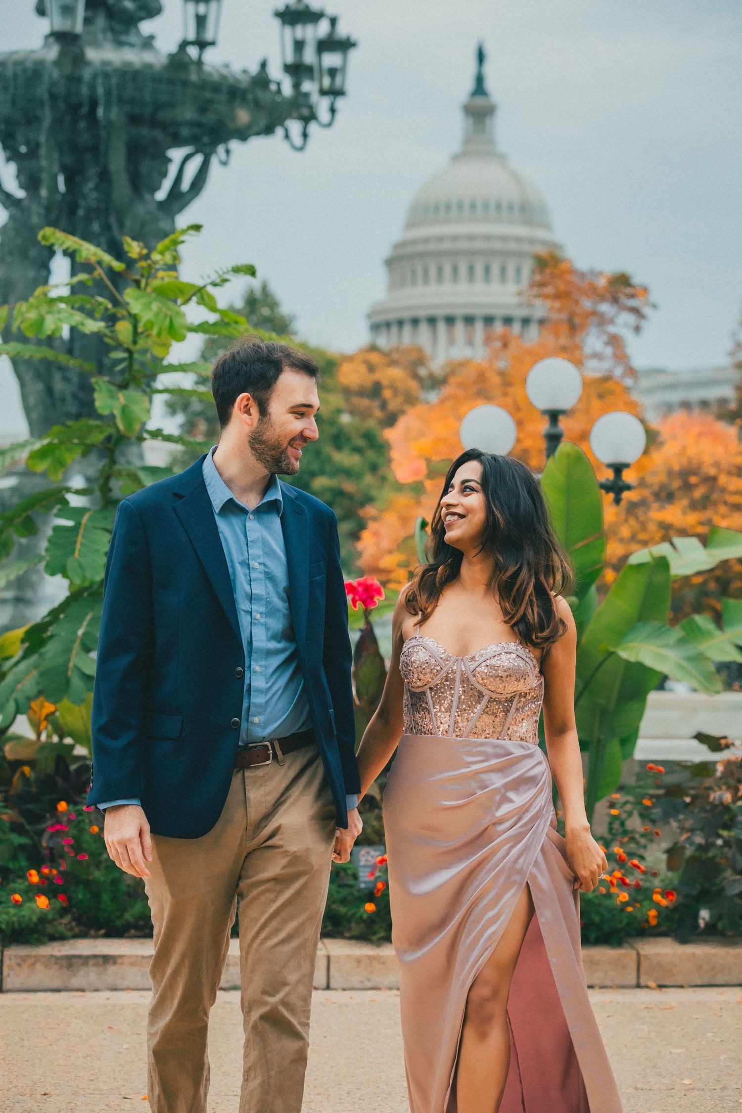 Smiling couple walking hand in hand through autumn foliage near the U.S. Capitol dome in Washington, DC, during an engagement photo shoot.