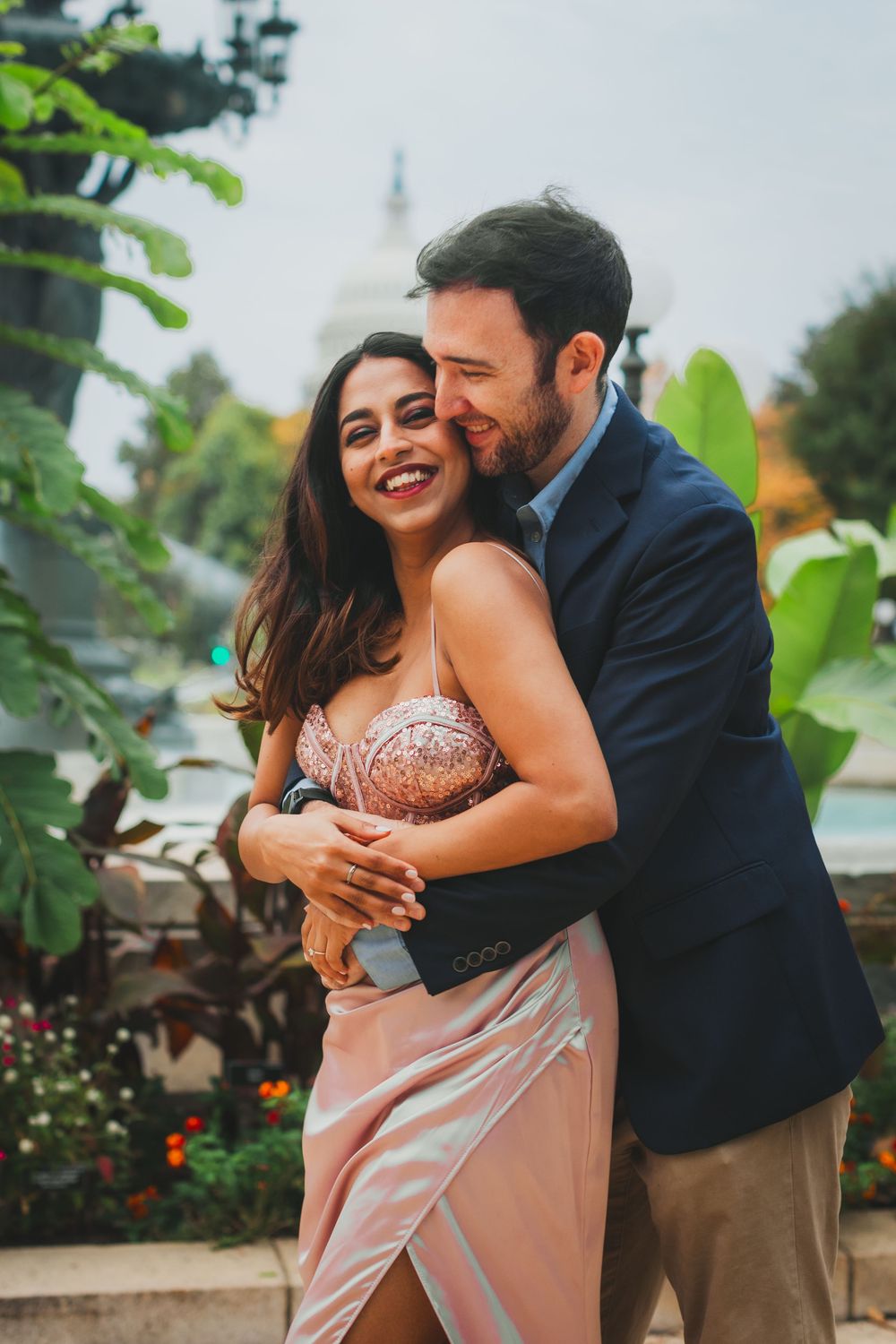 Smiling couple embracing in a lush garden, she in a blush satin dress and he in a navy blazer, with a domed building behind.