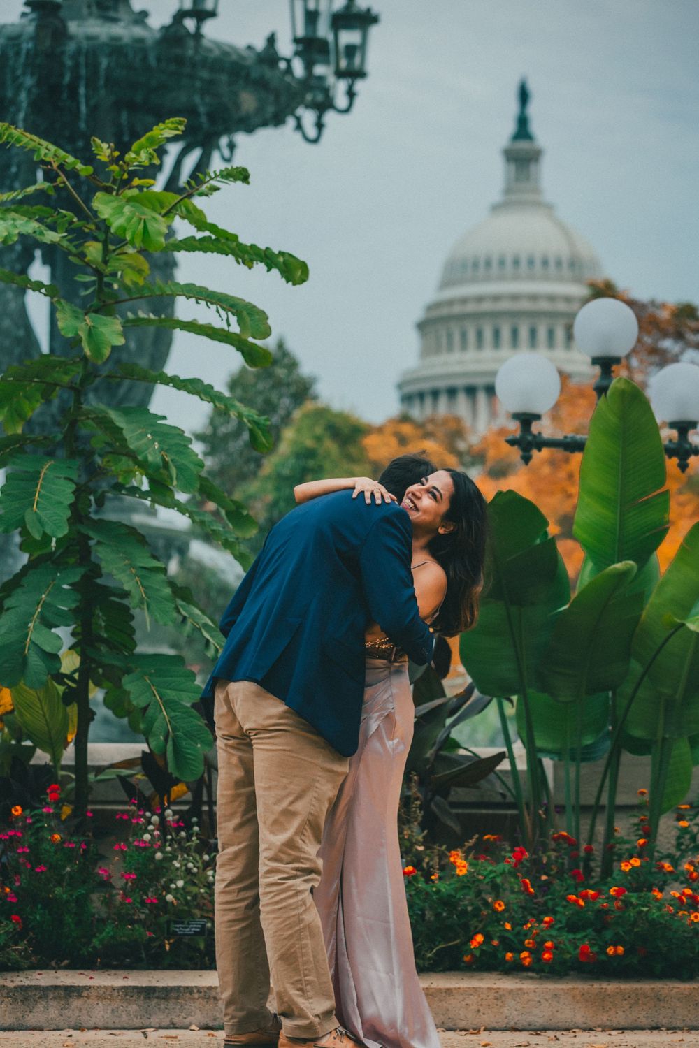 Couple embracing in a garden with tropical plants and autumn flowers, with the U.S. Capitol dome rising in the background.