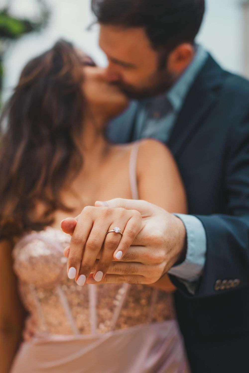 Couple kissing while holding hands in the foreground, showing a sparkling diamond engagement ring on her finger, she wears a pink dress.