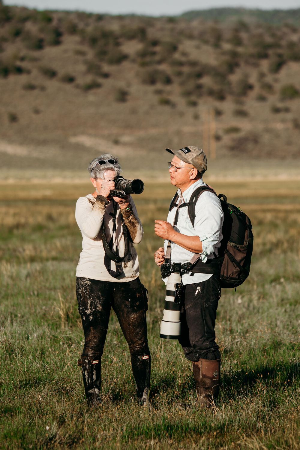 Two wildlife photographers with cameras and telephoto lenses stand in a grassy field, talking, with sagebrush-covered hills behind them.