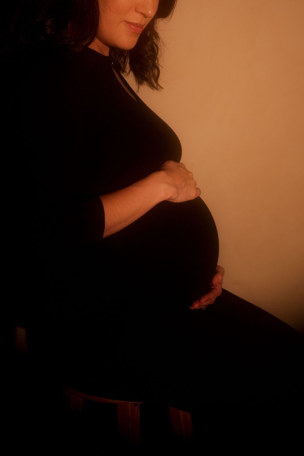 Pregnant woman in black dress cradling her baby bump in dramatic warm studio lighting.