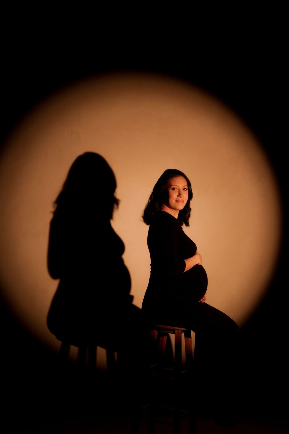 Pregnant woman sitting on stool in dramatic spotlight with shadow silhouette on warm background.