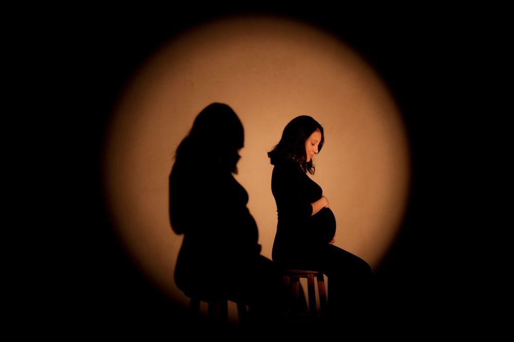 Dramatic maternity photo of pregnant woman with shadow silhouette against warm circular spotlight on dark background.