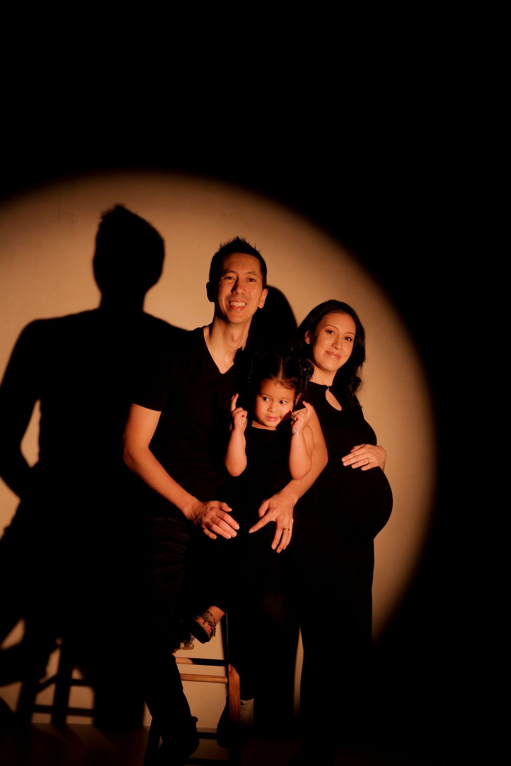 Family maternity photo with parents and child in black outfits against warm spotlight backdrop.