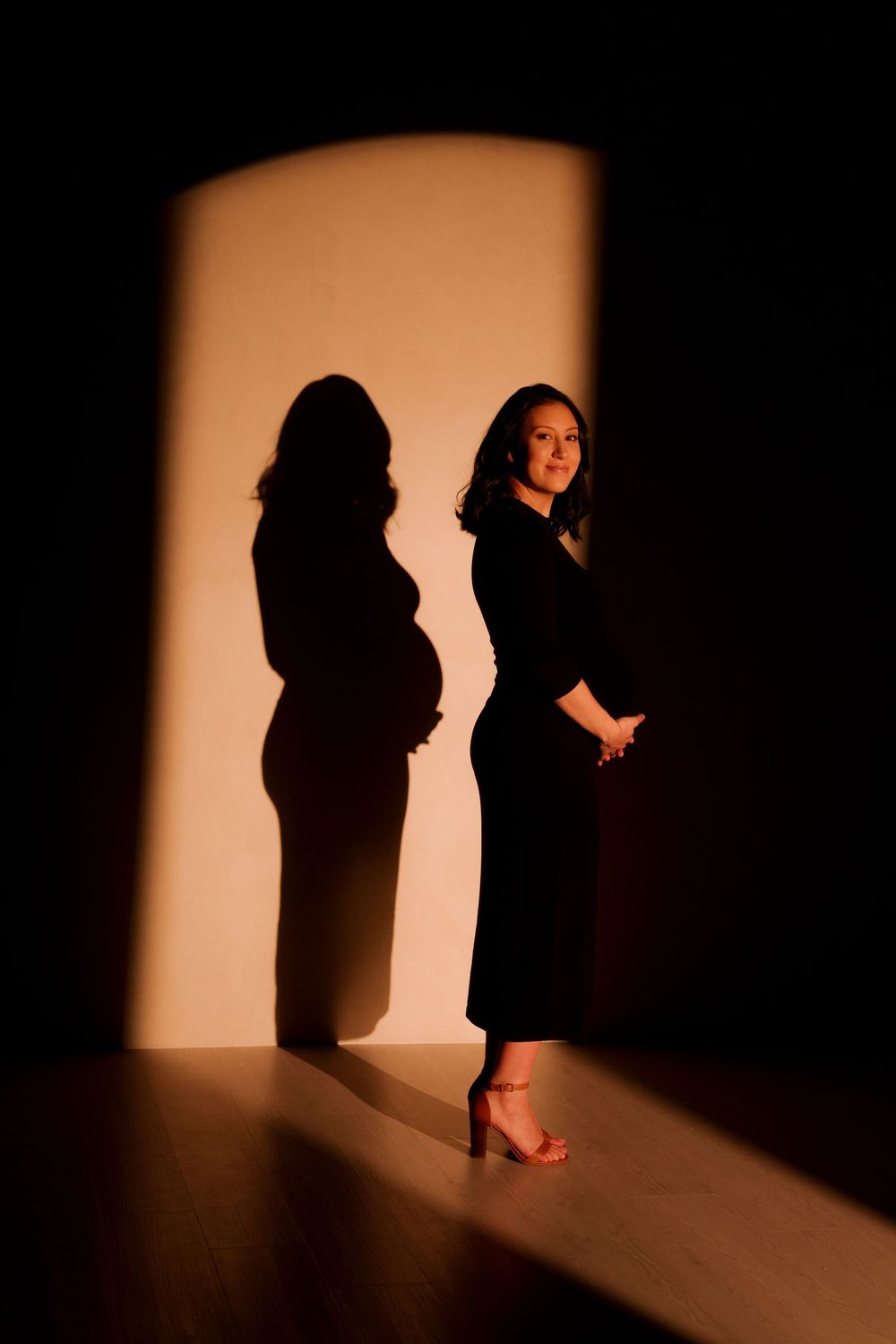 Pregnant woman in black dress with dramatic shadow silhouette on warm lit wall.