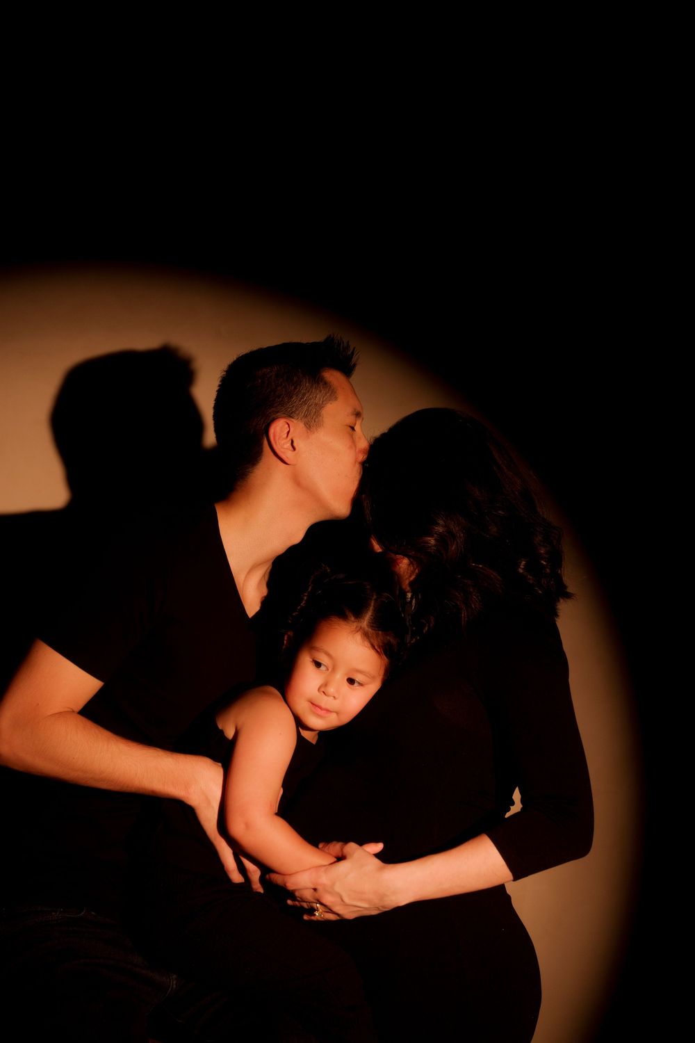 A couple in black clothing kiss tenderly while holding their young child in a dramatic spotlight.