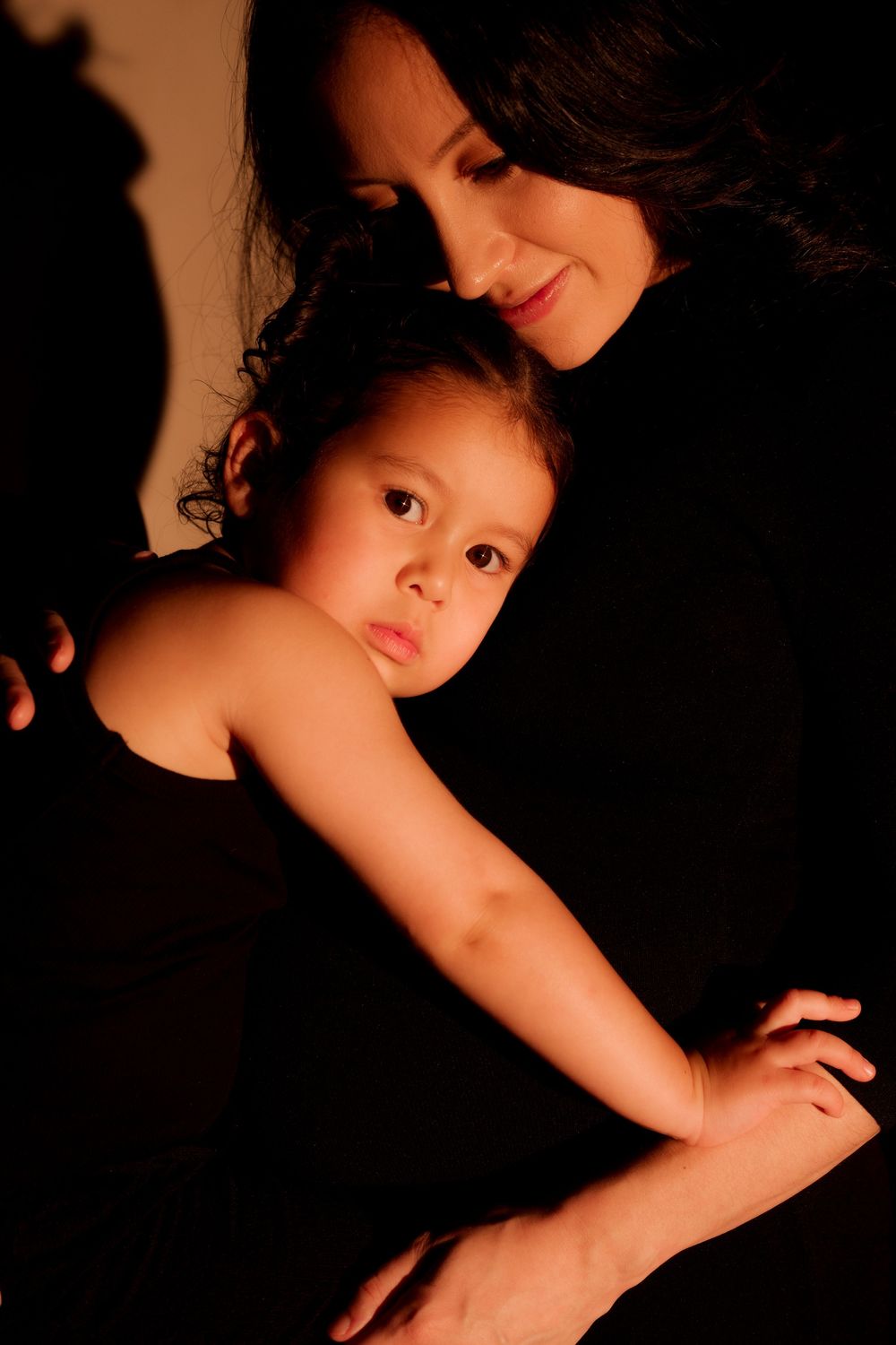 A woman in black tenderly embraces a young child in a warm, dimly lit portrait.