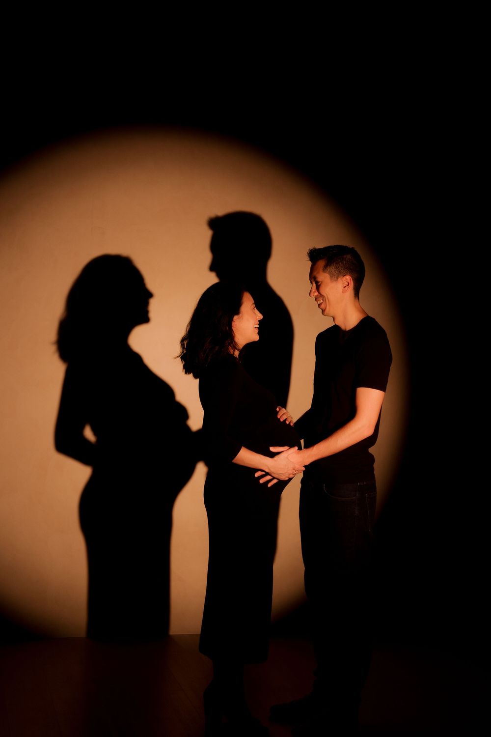Expectant couple standing together with dramatic shadows cast on warm-lit studio background.