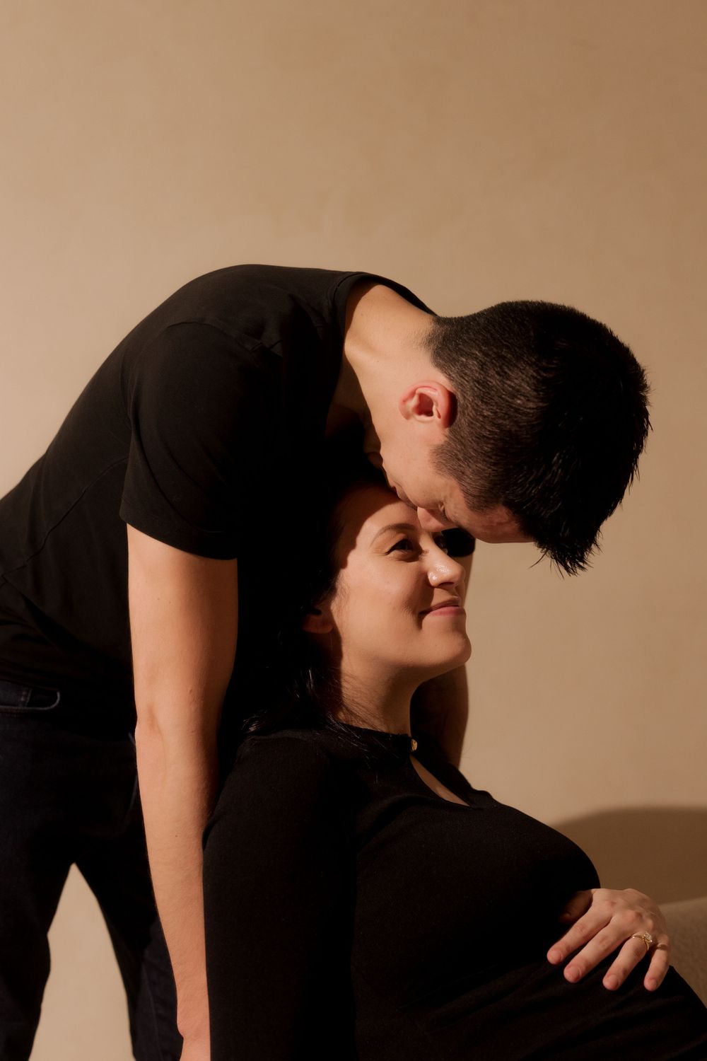 A man in black tenderly kisses a pregnant woman's head while gently touching her baby bump in a studio portrait.